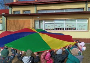 Grupa dzieci macha chustą animacyjną podczas zabawy na dworze.