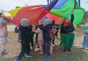 Dzieci unoszą i opuszczają dużą kolorową chustę podczas zabawy na dworze.