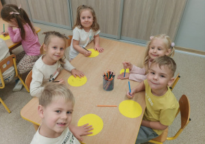 Dzieci uśmiechają się do zdjęcia siedząc przy stoliku i wykonując pracę