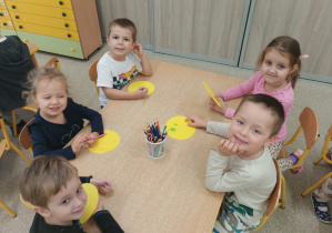 Dzieci uśmiechają się do zdjęcia siedząc przy stoliku i wykonując pracę