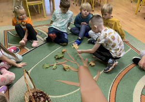 Dzieci siedzą na dywanie i wykonują z darów jesieni budowę drzewa