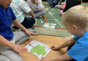 Dzieci siedzą na podłodze i układają puzzle z wizerunkiem drzewa