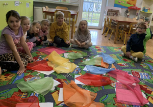 Dzieci podczas zajęć siedzą na dywanie i patrzą na ukryte pod chustkami karty