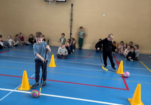 Dzieci toczą piłkę kijem hokejowym między pachołkami