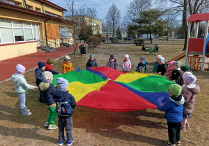 Przedszkolaki trzymają tęczową chustę i bawią się nią na dworze.