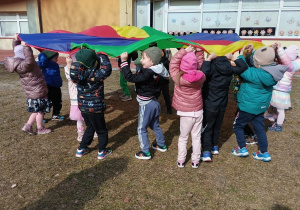 Dzieci stoją w kręgu i poruszają kolorową chustą na placu zabaw.